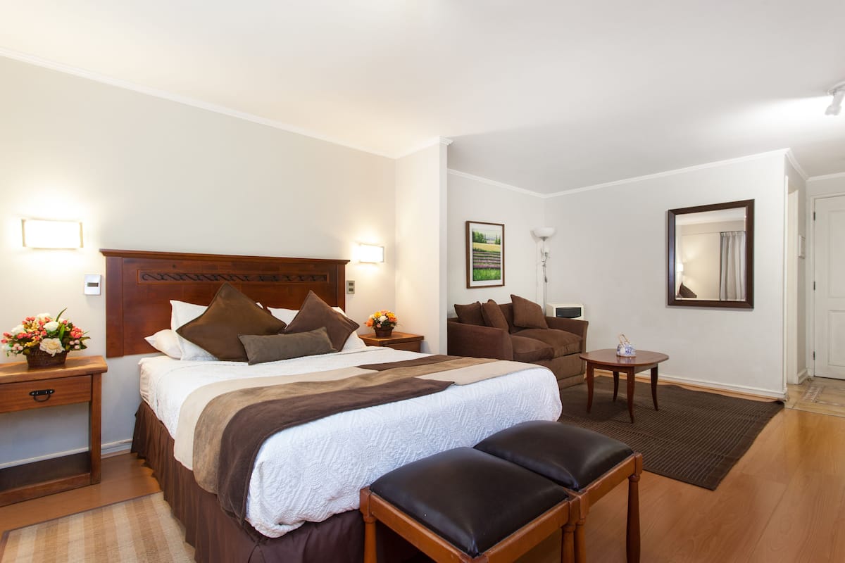 Una camera d'albergo con un letto grande, una testiera in legno, una zona salotto con divano e poltrona, un tavolino e uno specchio a parete.