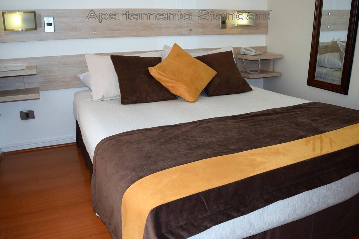 Una camera da letto ordinata con un letto, cuscini, comodini e uno specchio.