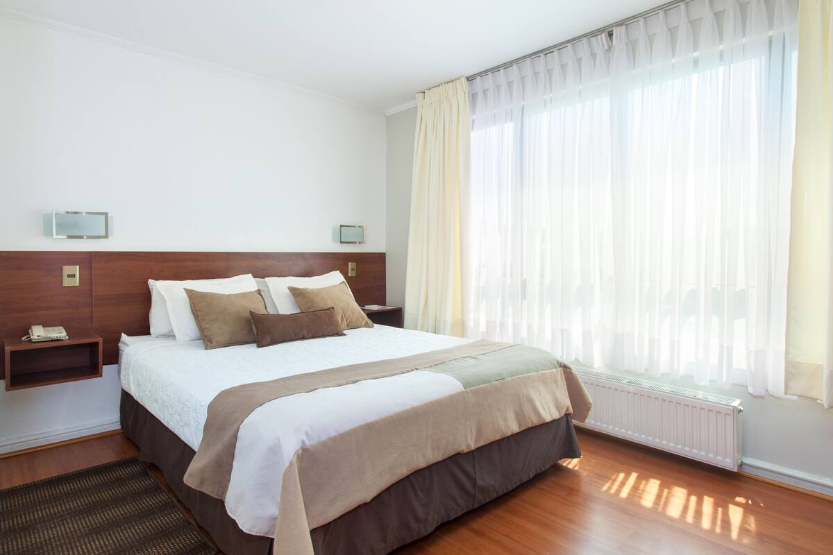 Camera d'albergo con un letto grande, una testata del letto in legno, un comodino con una lampada, una finestra con tende trasparenti e una TV a parete.