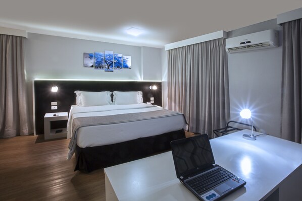 Una habitación de hotel con una cama, mesitas de noche, un escritorio con una computadora portátil, un equipo de aire acondicionado y un cuadro colgado en la pared.