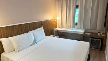 Quarto de hotel com cama grande, escrivaninha, cadeira, abajur e vista para o exterior.