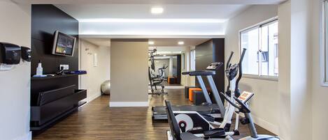 A modern gym with cardio machines, a wall-mounted TV, and a black reception desk.