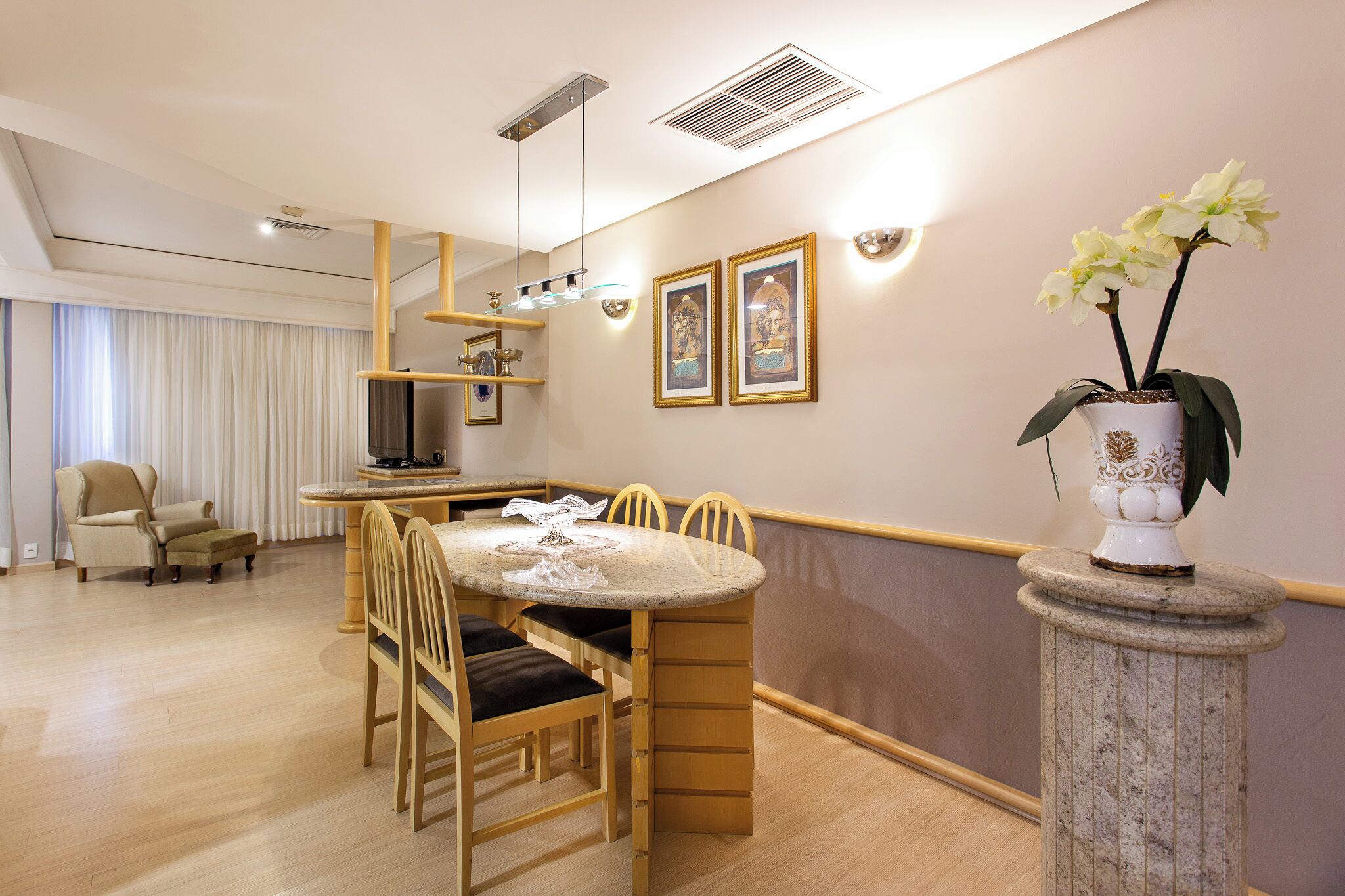 A dining area with a round table, chairs, a vase with flowers, and framed pictures on the wall.