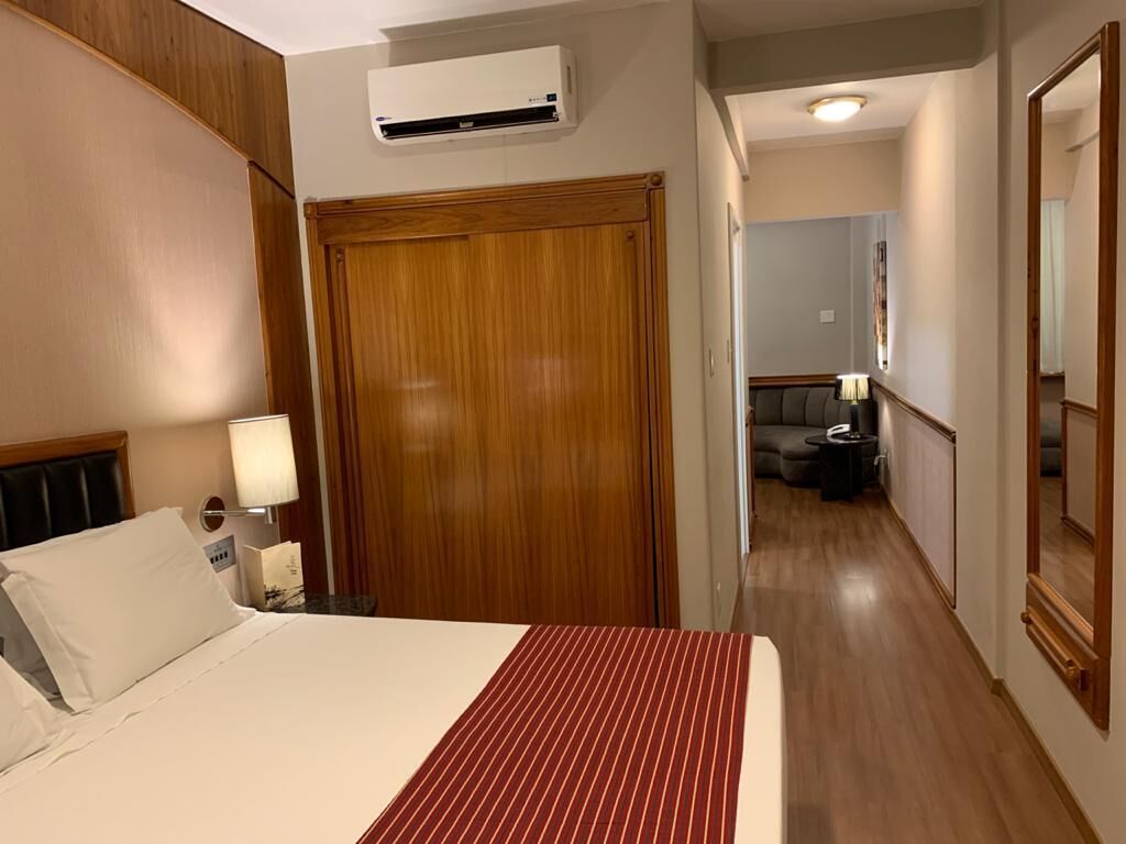 Habitación de hotel con cama, armario de madera, mesita de noche con lámpara y un pasillo que conduce a otra habitación.