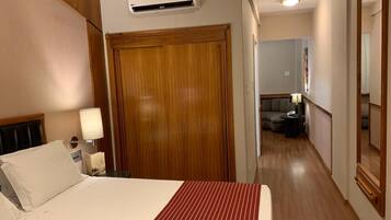 Habitación de hotel con cama, armario de madera, mesita de noche con lámpara y un pasillo que conduce a otra habitación.
