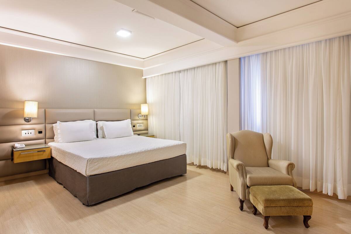 A hotel room with a large bed, bedside lamps, a chair, and a window with curtains.