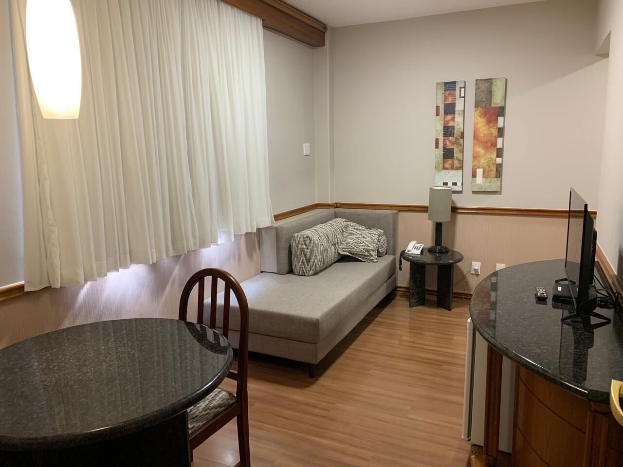 A hotel room with a sofa, a dining table, a chair, a lamp, and artwork on the wall.