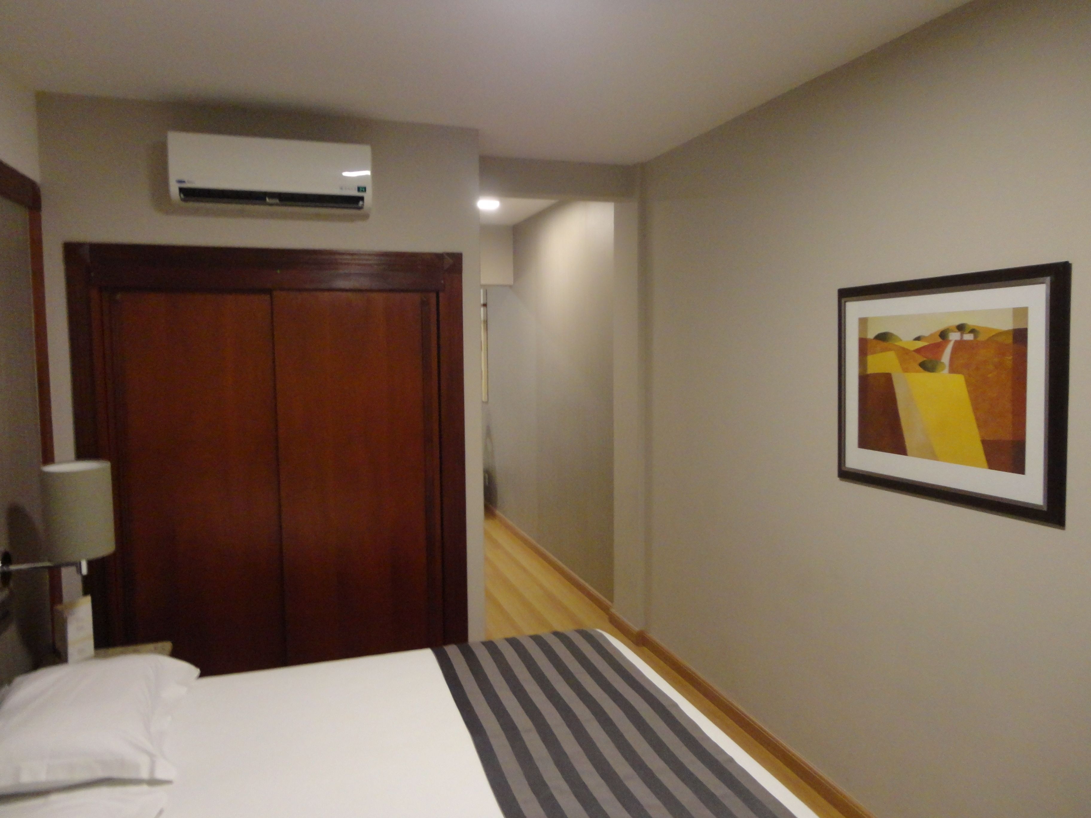 A hotel room with a bed, a wooden wardrobe, an air conditioning unit, a lamp, and a framed abstract painting on the wall.