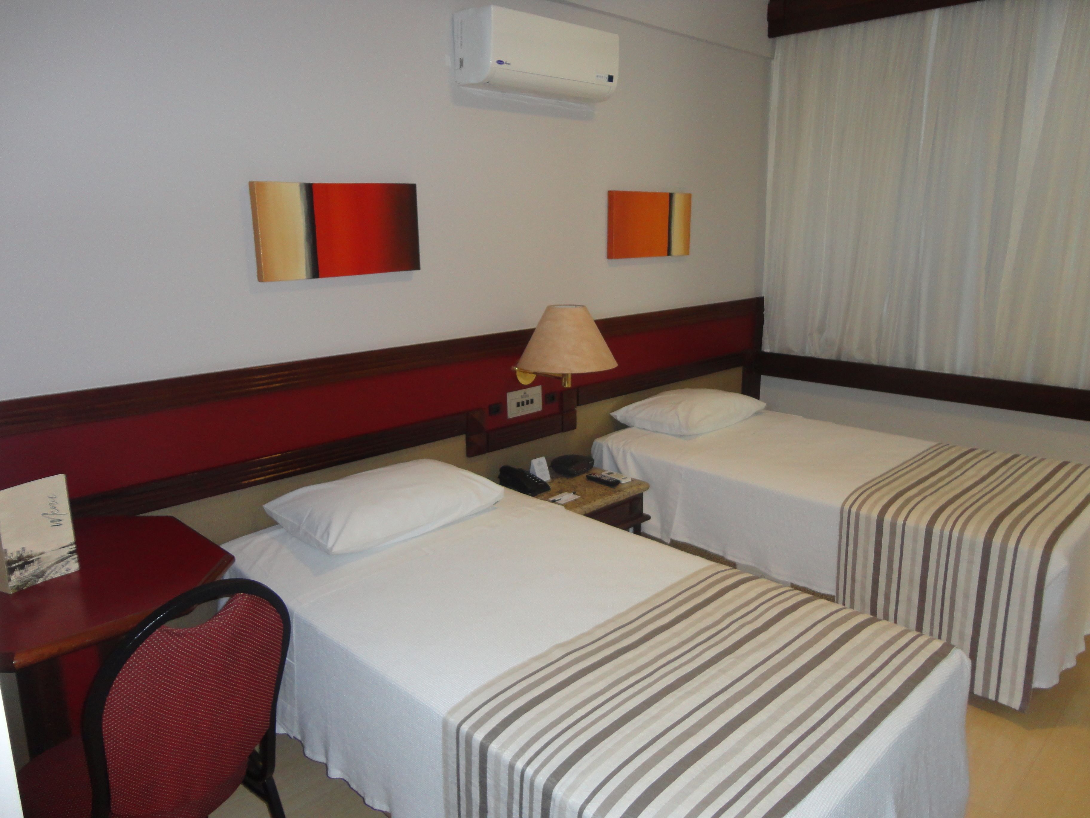 Habitación de hotel con dos camas individuales, un escritorio, una silla, una lámpara y dos cuadros colgados en la pared.
