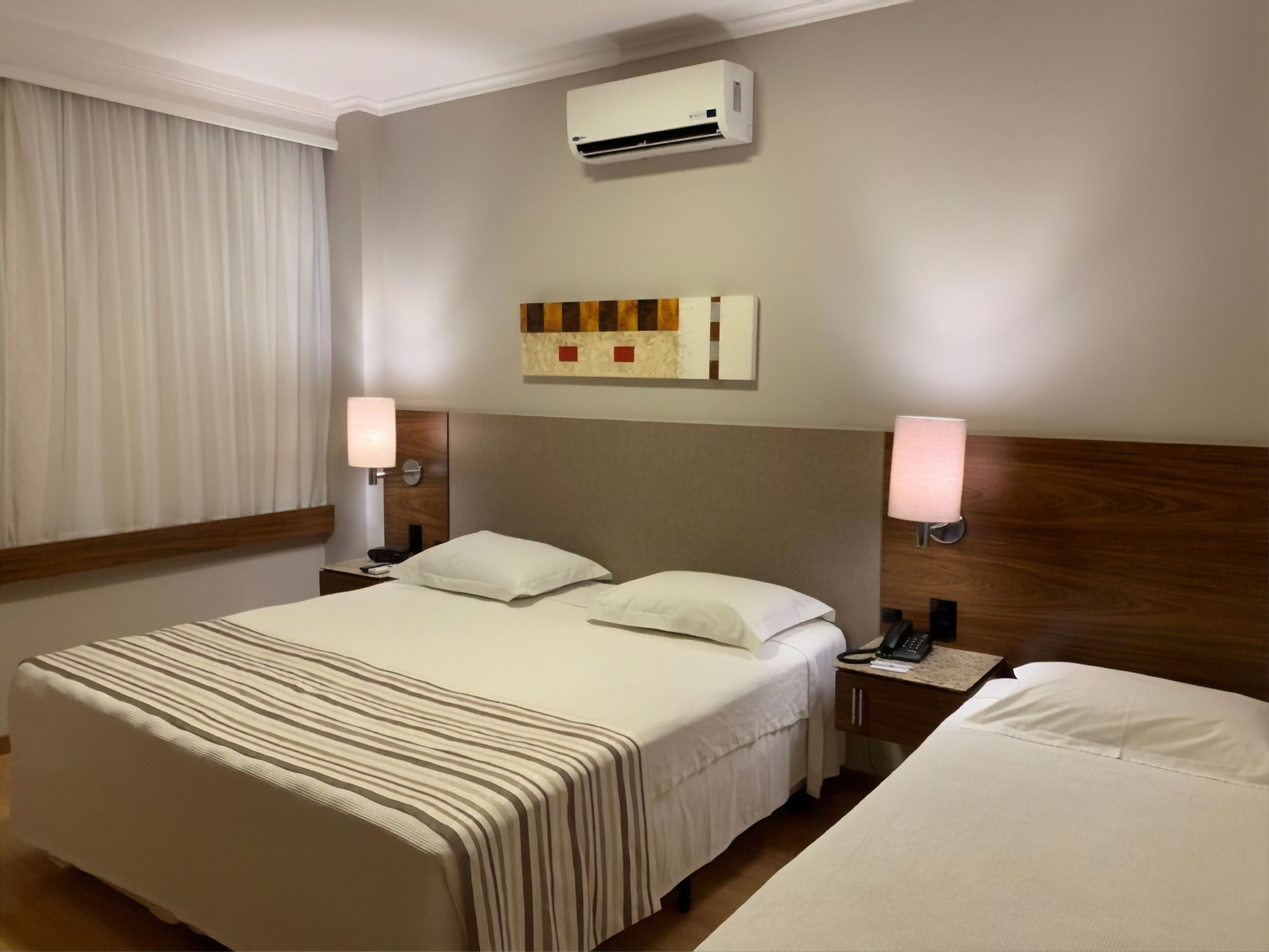 Quarto de hotel com uma cama grande, luminárias de cabeceira, telefone e ar condicionado na parede.