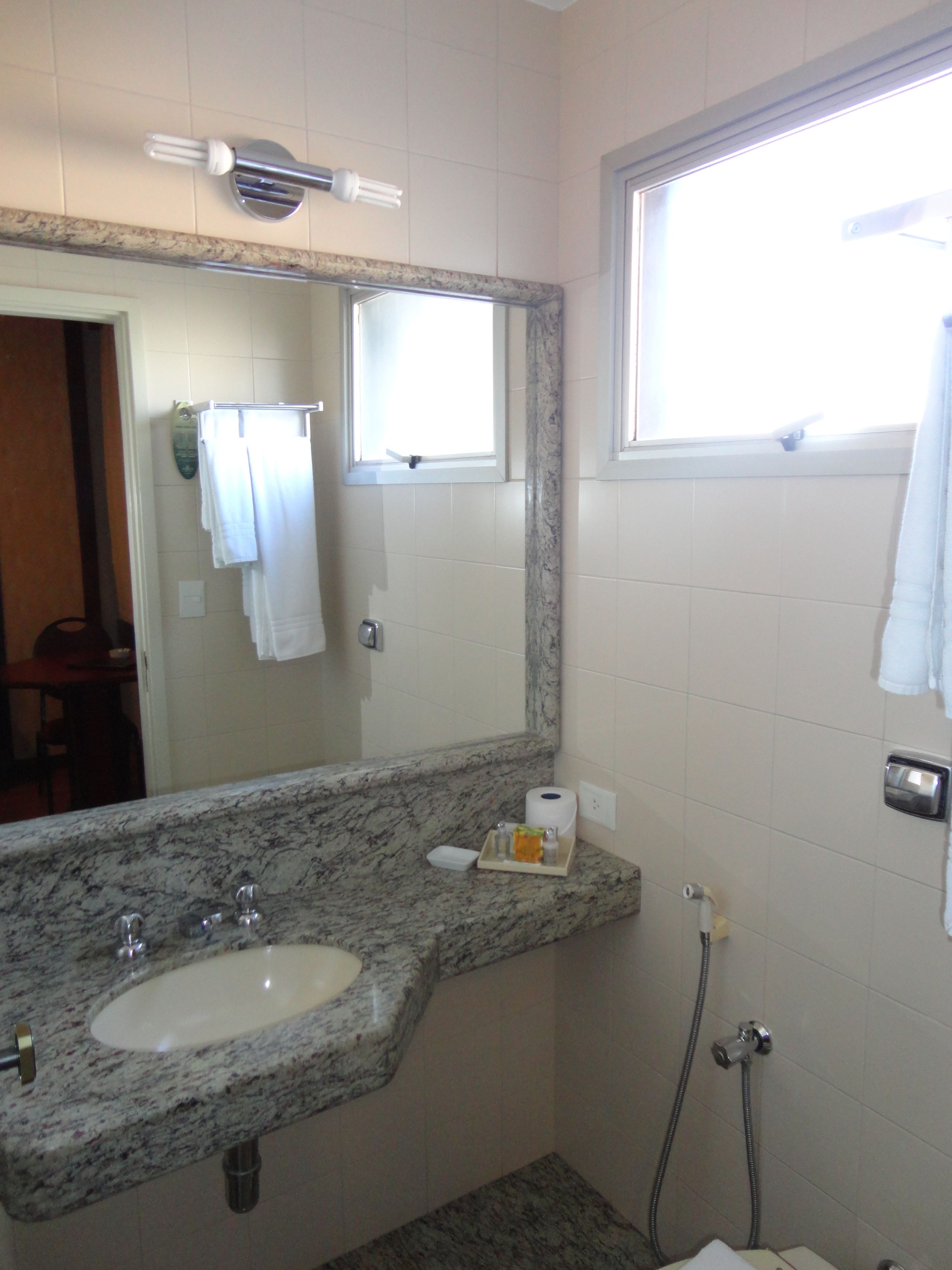A bathroom with a granite countertop sink, a mirror, a towel rack, a window, and a shower.