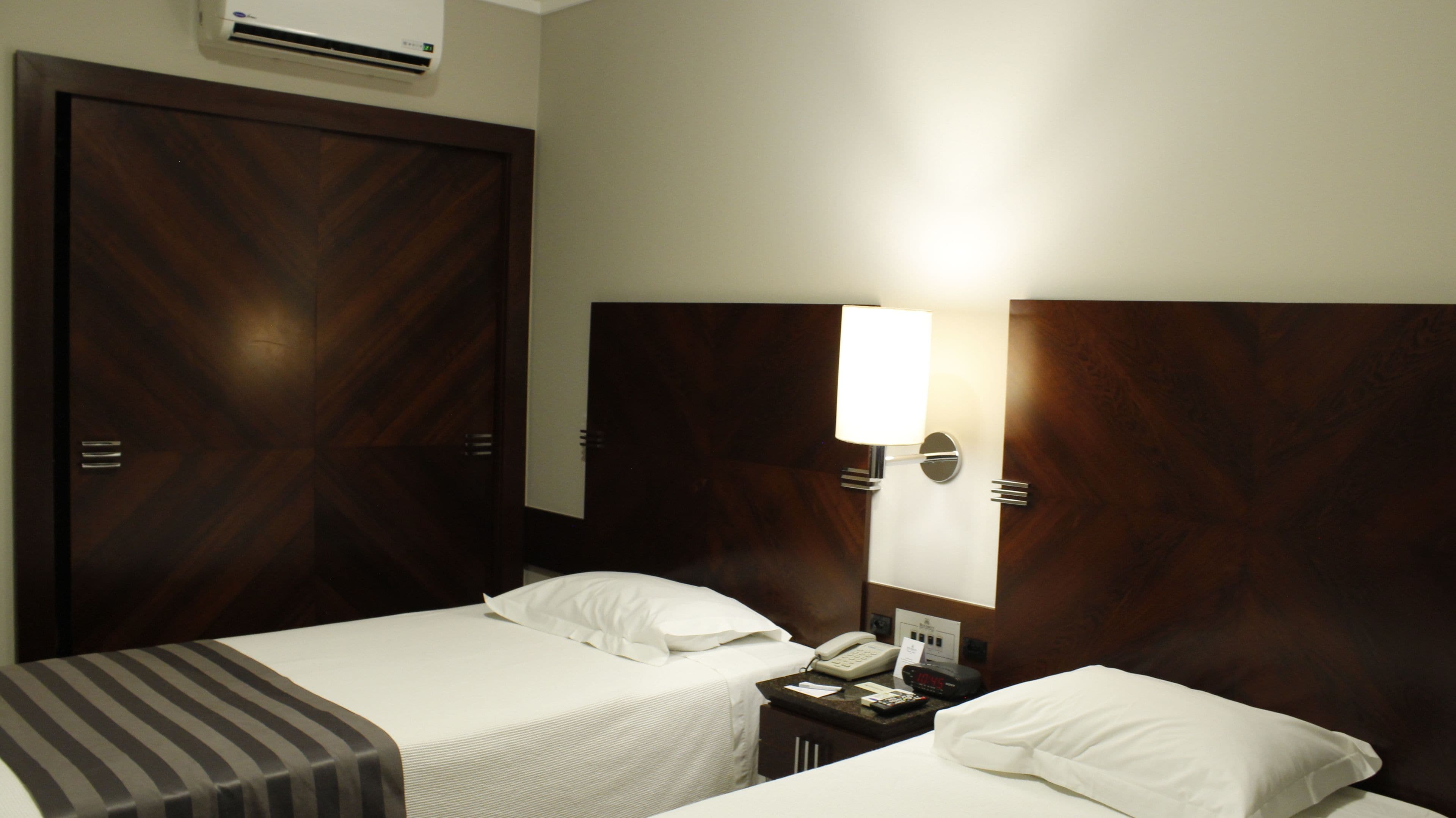 A hotel room with two beds, a bedside table with a phone and a clock, and a wall-mounted lamp.