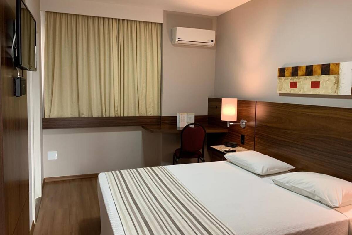 A hotel room with a bed, a desk, a chair, a TV, an air conditioner, and a window with curtains.