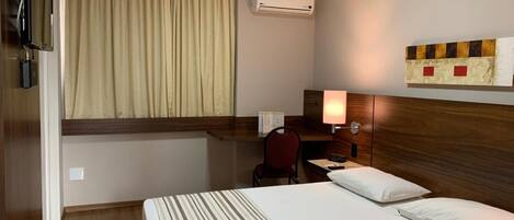 Una camera d'albergo con un letto, una scrivania, una sedia, una televisione, un condizionatore d'aria e una finestra con tende.