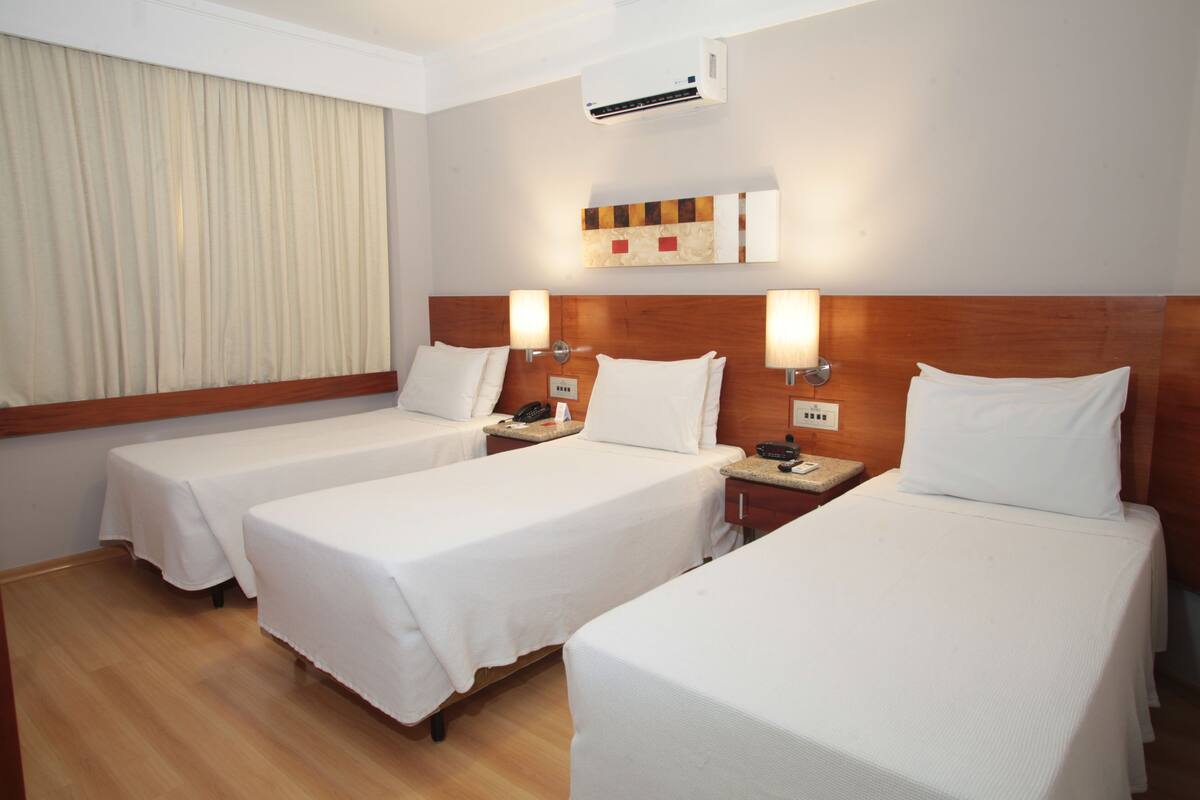 A hotel room with three single beds, a wooden headboard, a wall-mounted air conditioner, and a bedside table with a lamp.