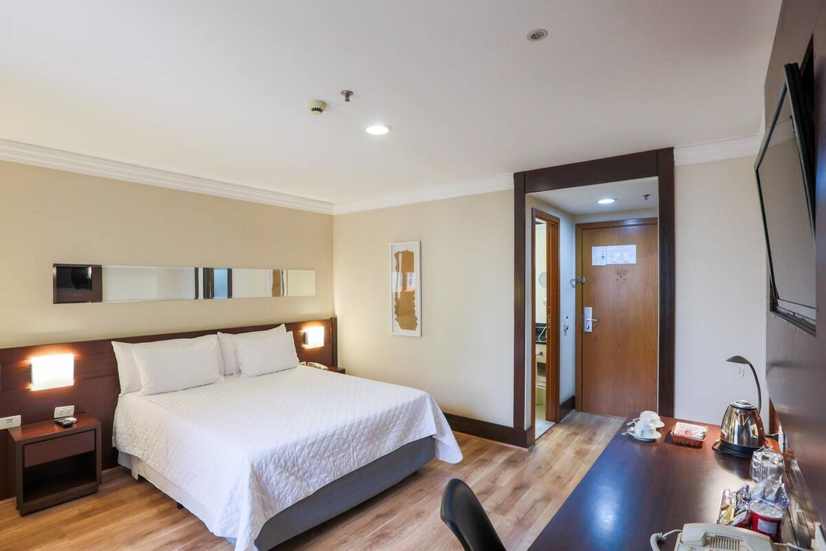 Habitación de hotel con cama, mesita de noche, escritorio con silla, televisor y puerta que da a otra habitación.