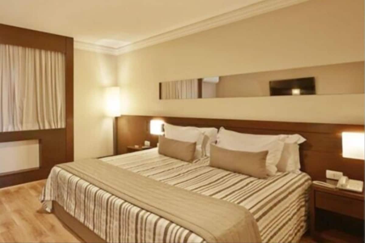 Una habitación de hotel con una cama grande, dos mesitas de noche con lámparas, un cabecero de madera y un ventanal con cortinas.