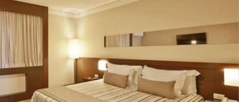 Una habitación de hotel con una cama grande, dos mesitas de noche con lámparas, un cabecero de madera y un ventanal con cortinas.