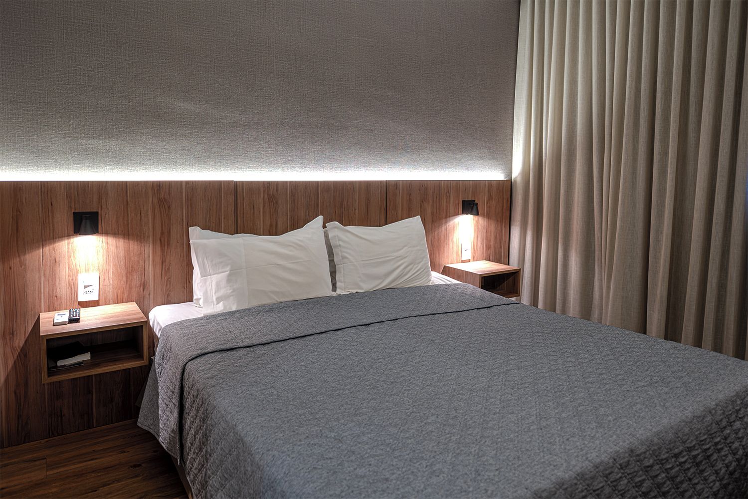 A hotel room with a bed, two bedside tables, a nightstand, a wall-mounted lamp, and a curtain.