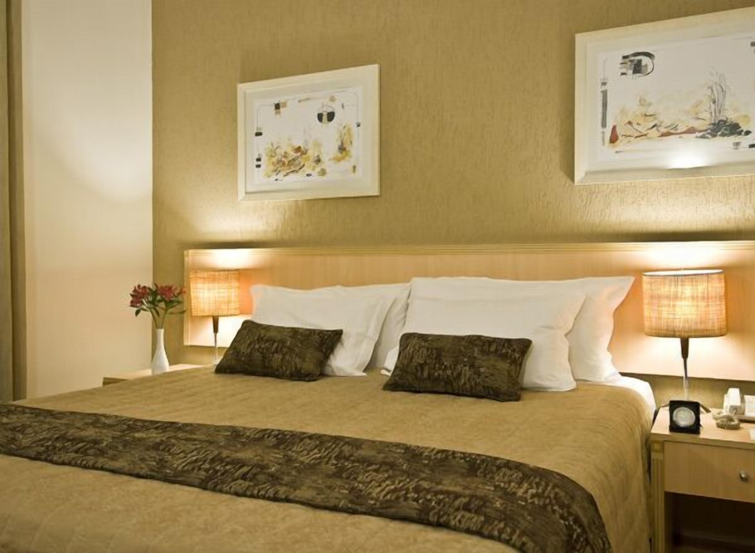Una habitación de hotel con una cama grande, dos lámparas de noche, una mesita de noche y dos cuadros en la pared.