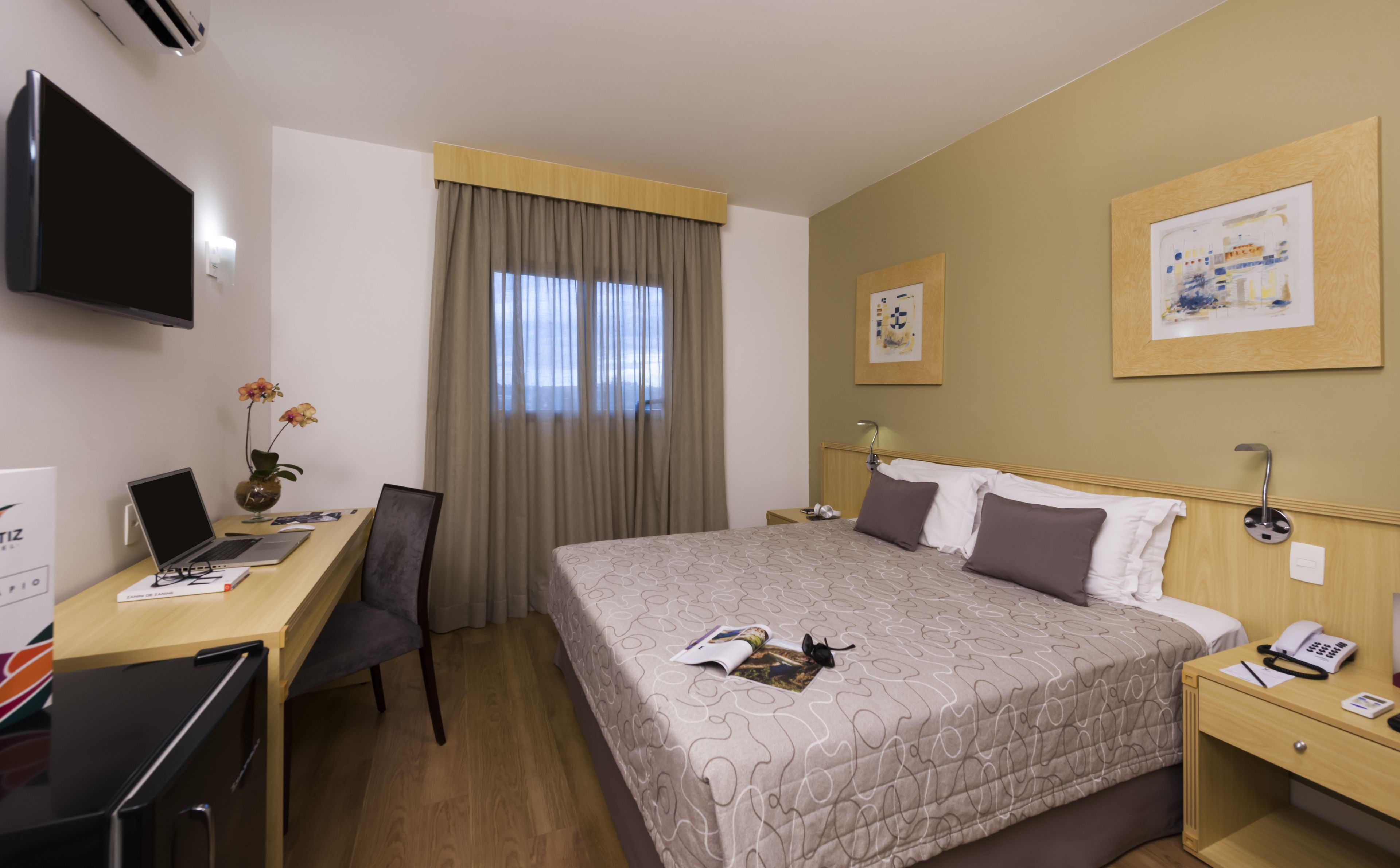 A hotel room with a bed, a desk with a laptop, a TV, and a window with curtains.