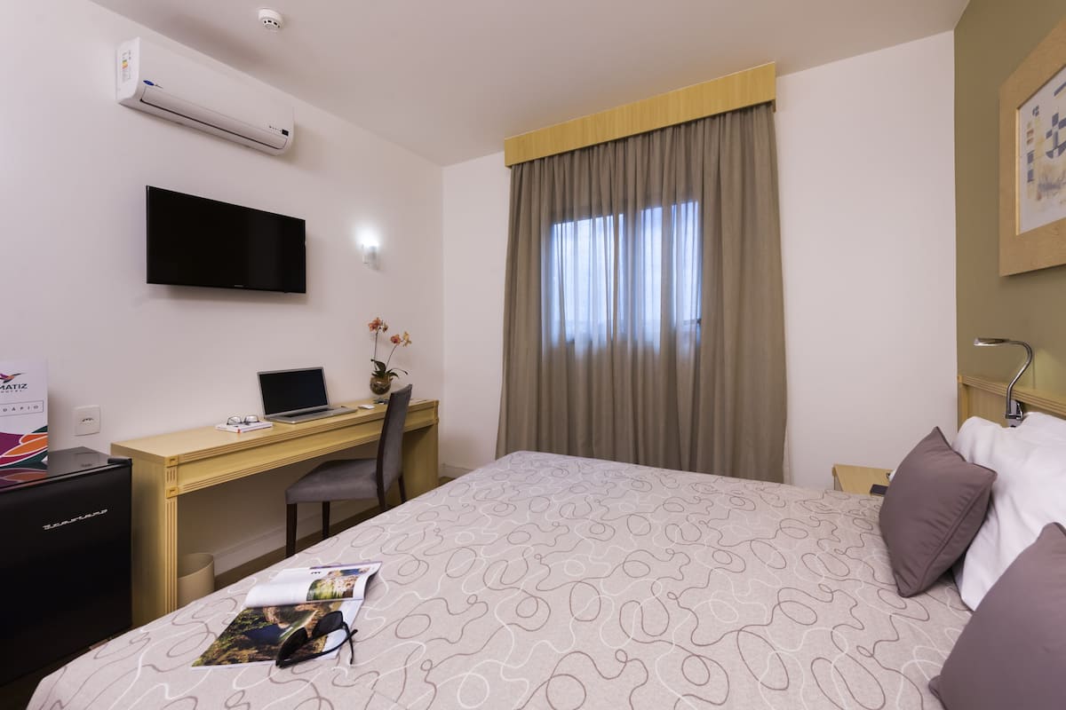 A hotel room with a bed, a desk with a laptop, a TV, and an air conditioner.