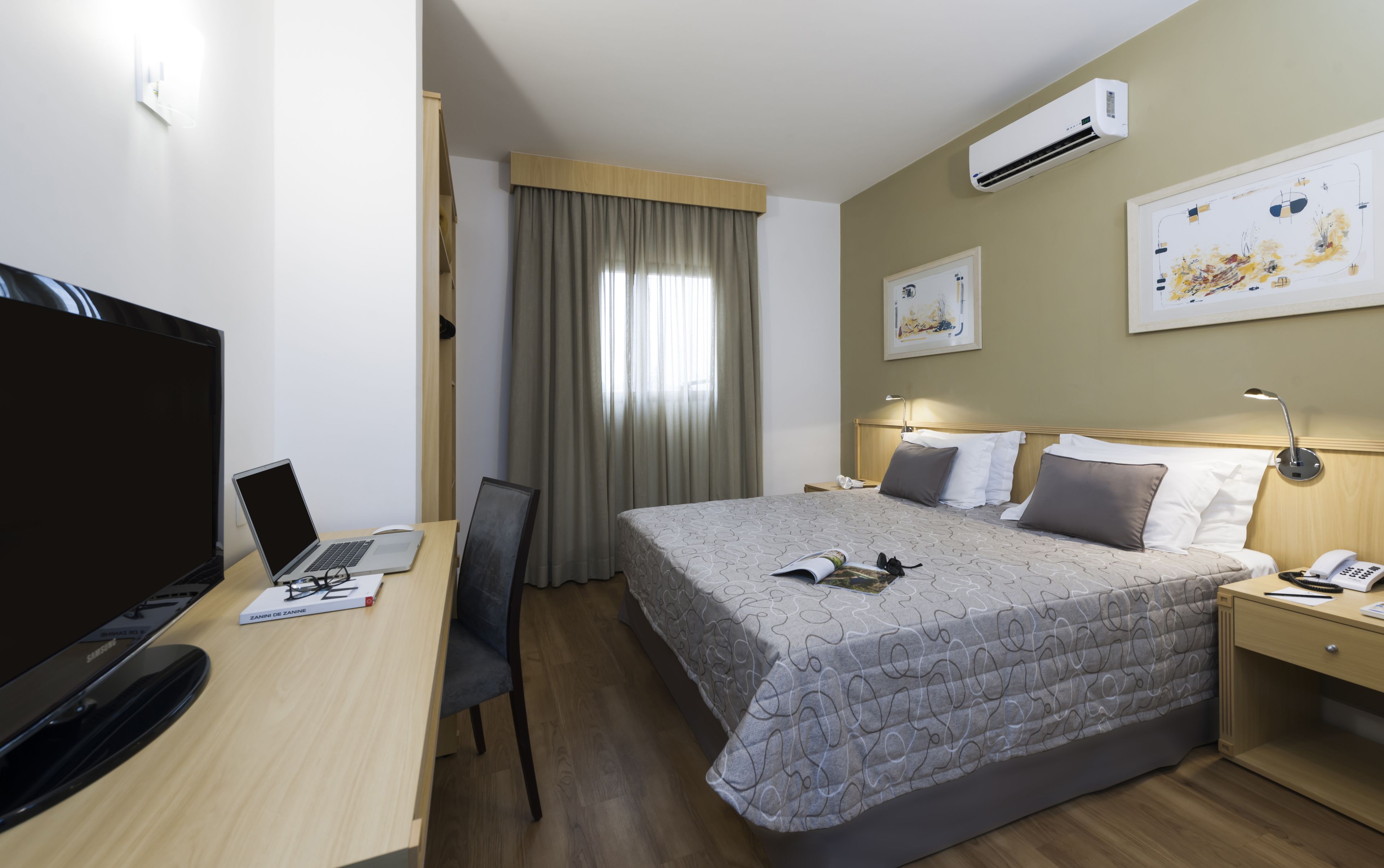 Una habitación de hotel con una cama, un escritorio con una computadora portátil, un televisor y un equipo de aire acondicionado.