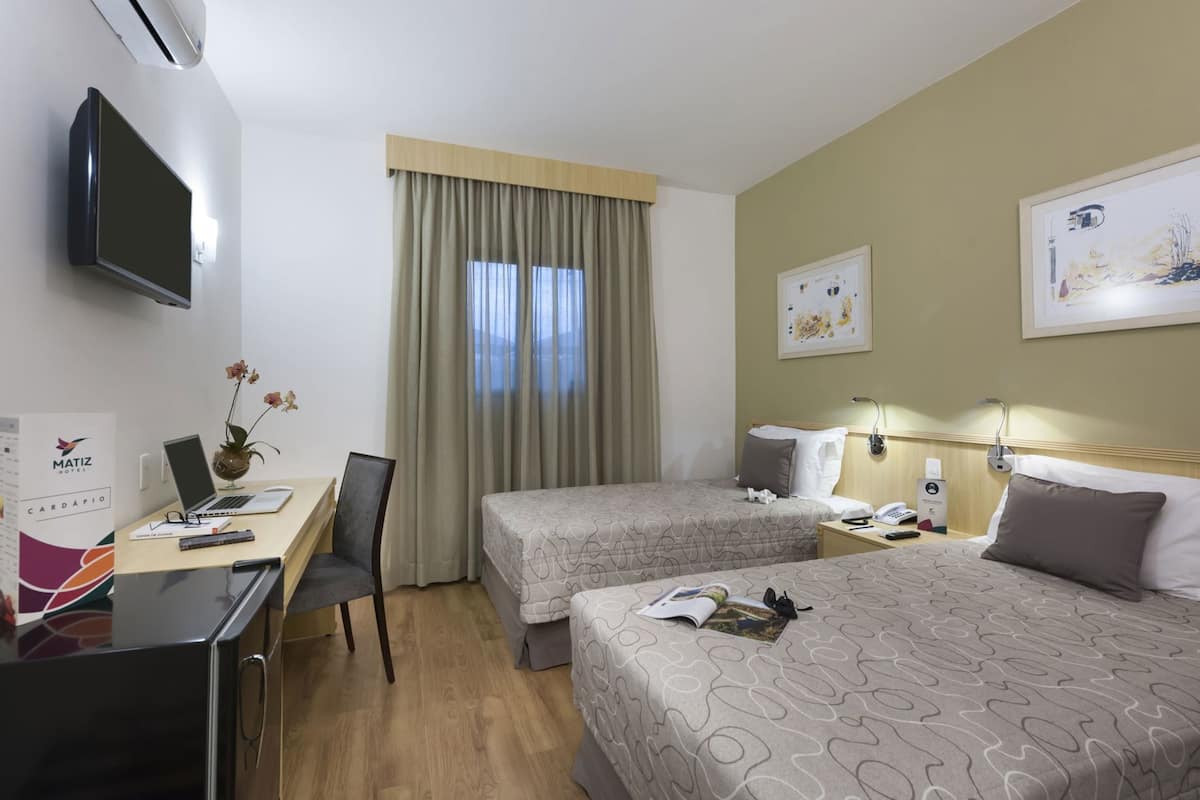 A hotel room with two beds, a desk with a laptop, a chair, a TV, and a window with curtains.