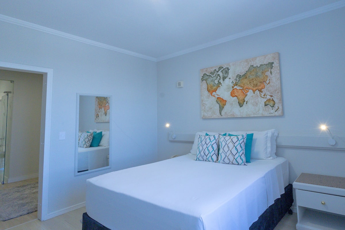 A bedroom with a bed, a nightstand, a wall-mounted lamp, a world map artwork, and a mirror.