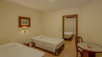 A hotel room with two single beds, a small table with a chair, a wall-mounted lamp, and a framed picture.