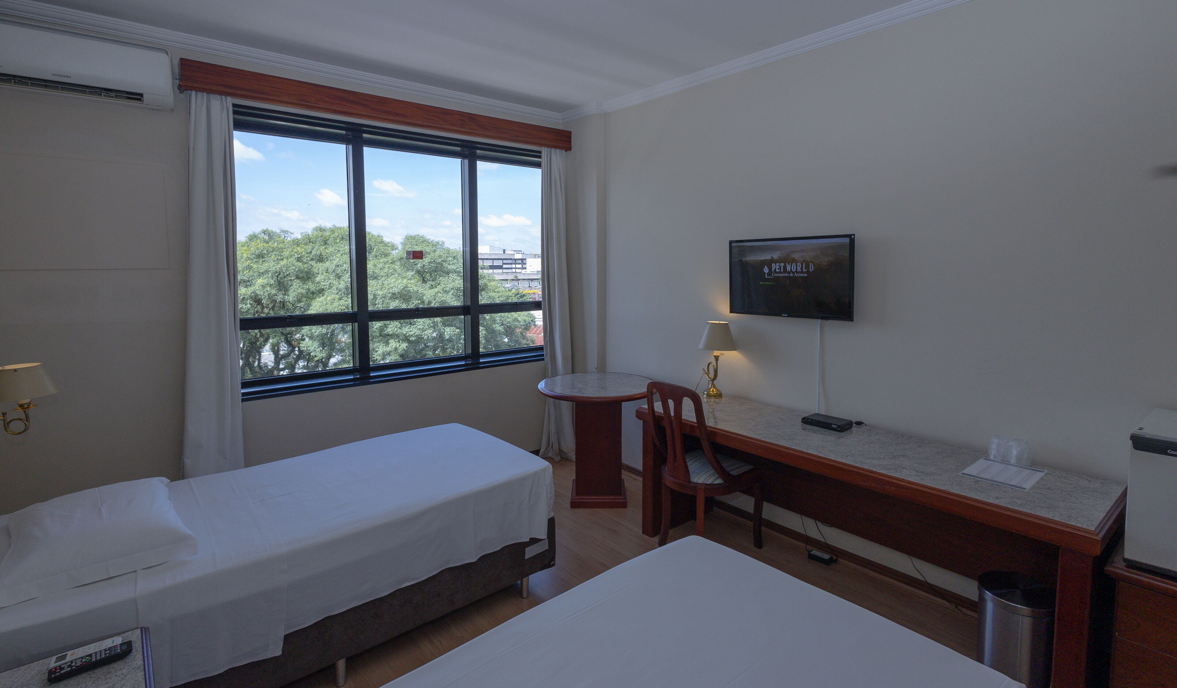 Habitación de hotel con dos camas individuales, un escritorio con silla, televisor y ventana con vistas a árboles y edificios.