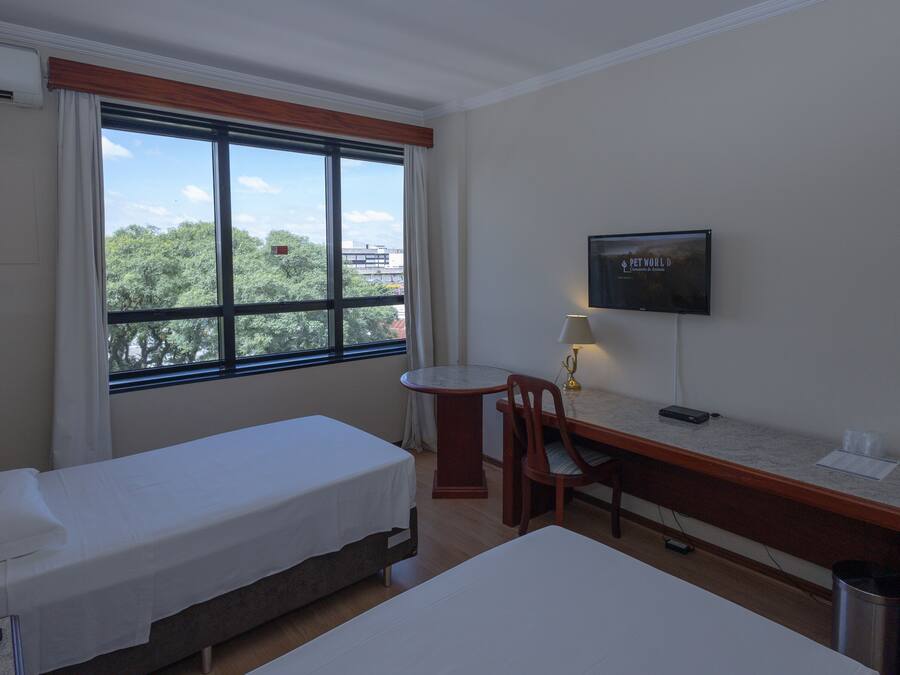 Habitación de hotel con dos camas individuales, un escritorio con silla, televisor y ventana con vistas a árboles y edificios.