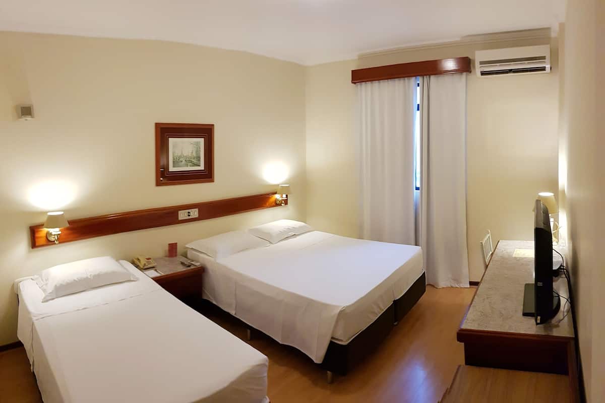 A hotel room with two single beds, a wooden headboard, a bedside table with a lamp, a framed picture on the wall, and an air conditioning unit.