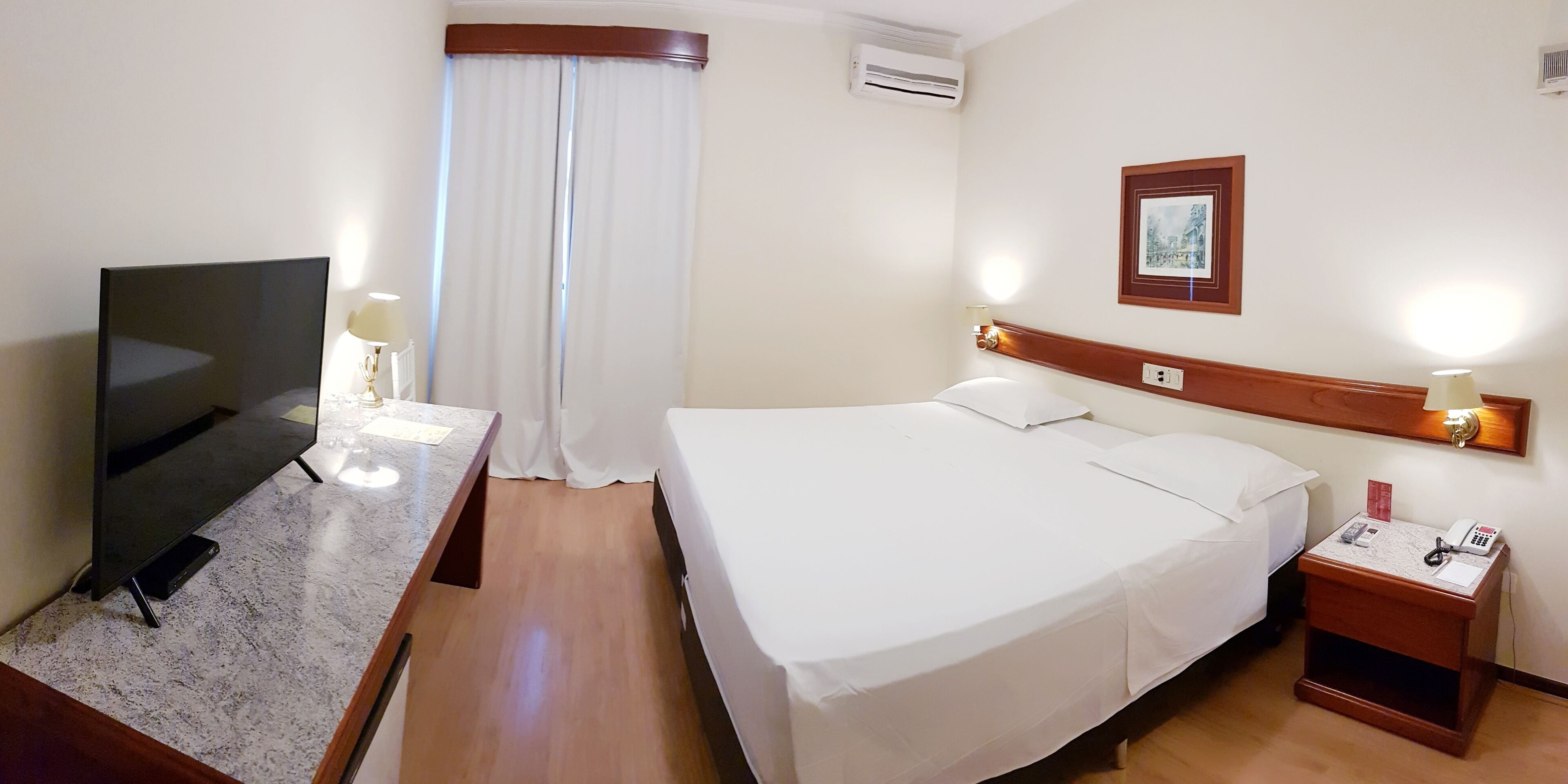 A hotel room with a bed, a desk with a TV, a nightstand, and a wall-mounted light fixture.