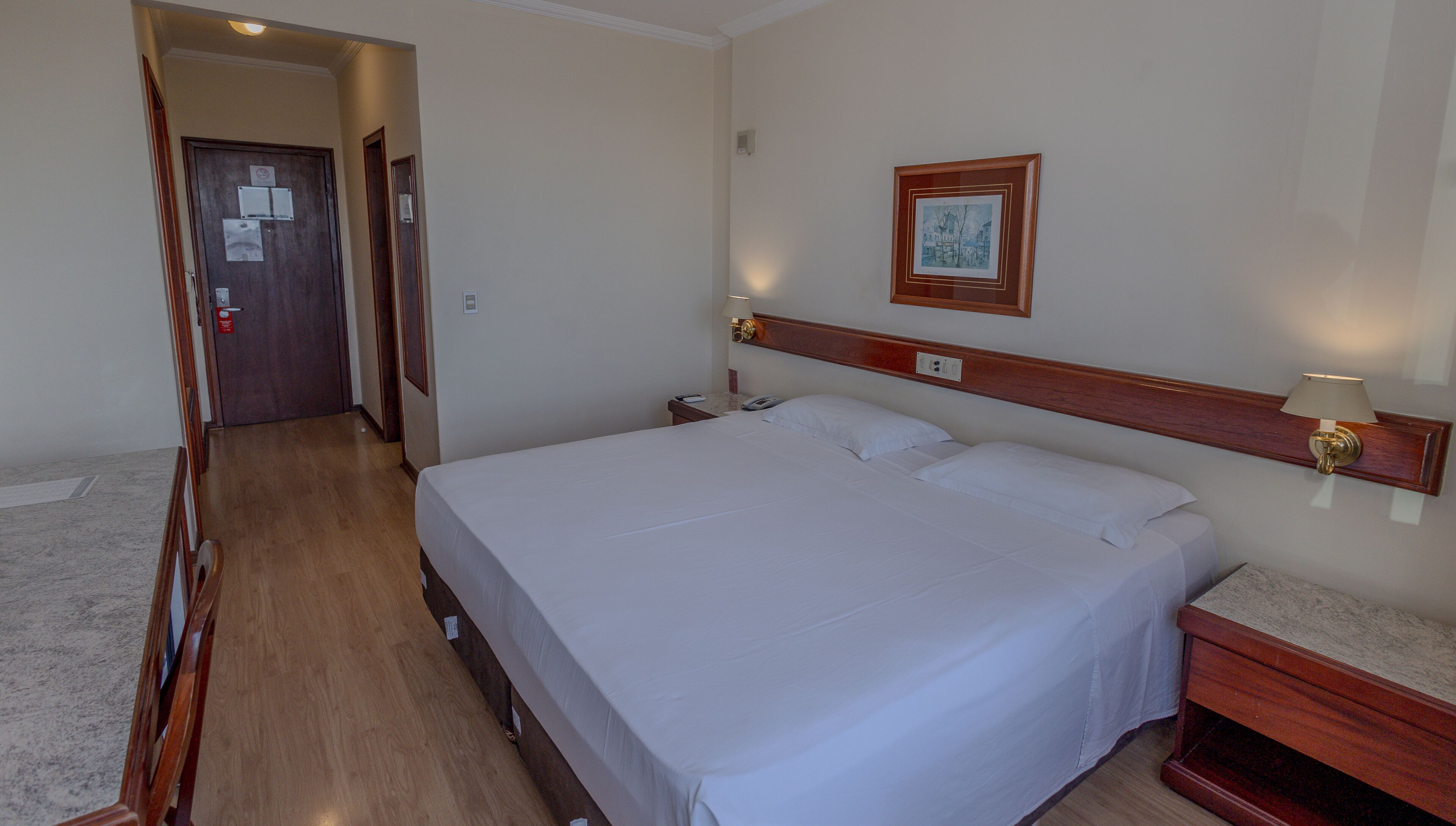 Une chambre d’hôtel avec un grand lit, une table de chevet, une commode, une lampe fixée au mur et une porte avec une pancarte.