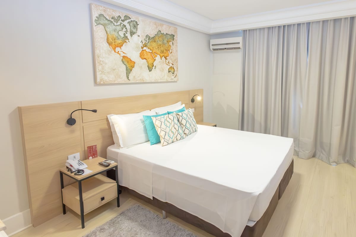 A hotel room with a large bed, a nightstand with a phone and remote, a world map artwork, an air conditioning unit, and a window with curtains.