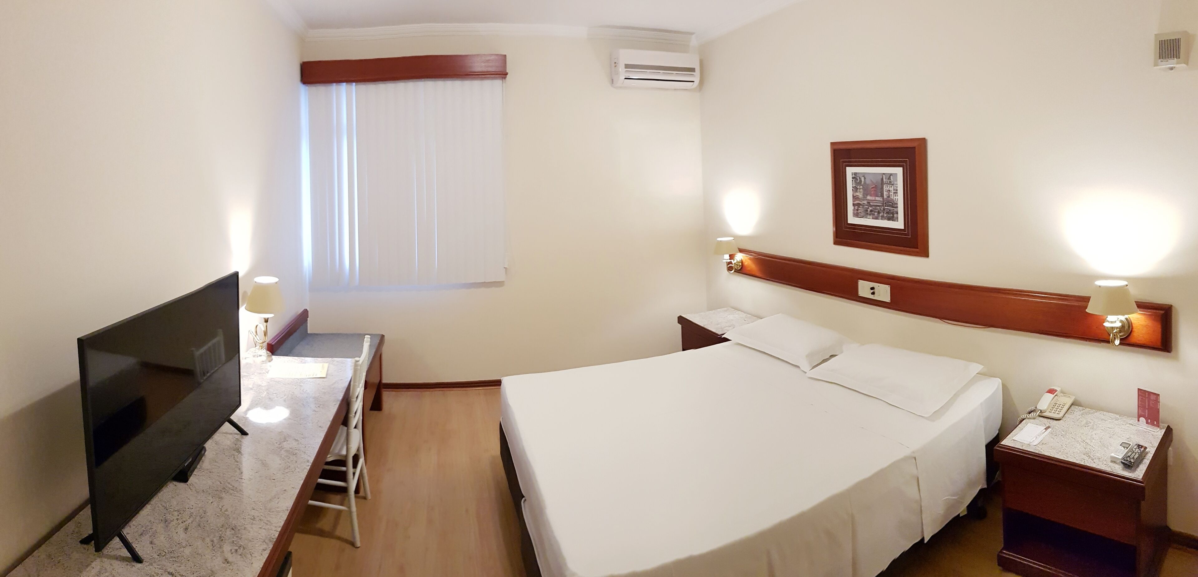 Una habitación de hotel con una cama, un escritorio con una silla, un televisor, una mesita de noche con una lámpara y un marco con un cuadro en la pared.