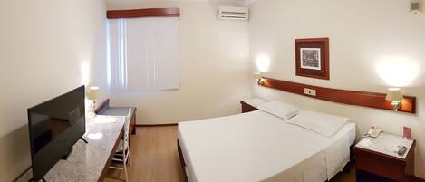 Una habitación de hotel con una cama, un escritorio con una silla, un televisor, una mesita de noche con una lámpara y un marco con un cuadro en la pared.