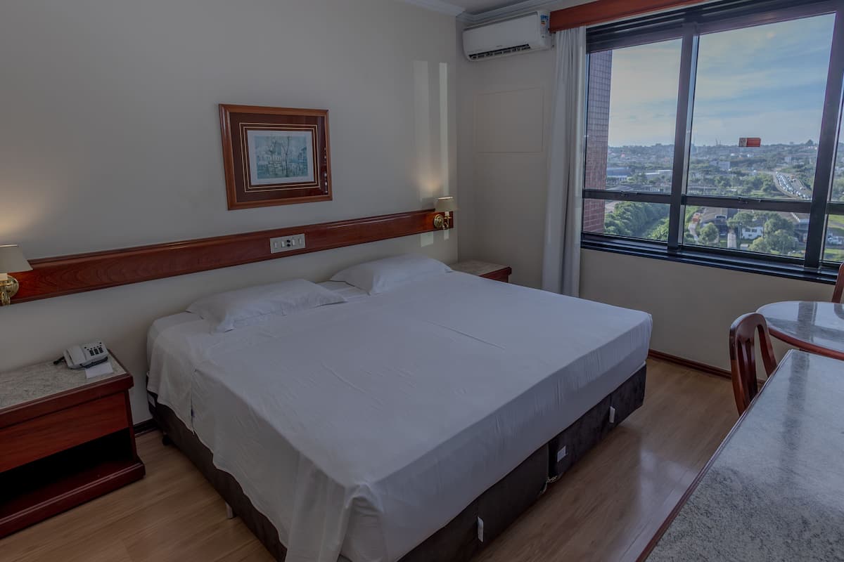 A hotel room with a bed, bedside table, lamp, and a view of a cityscape through a large window.