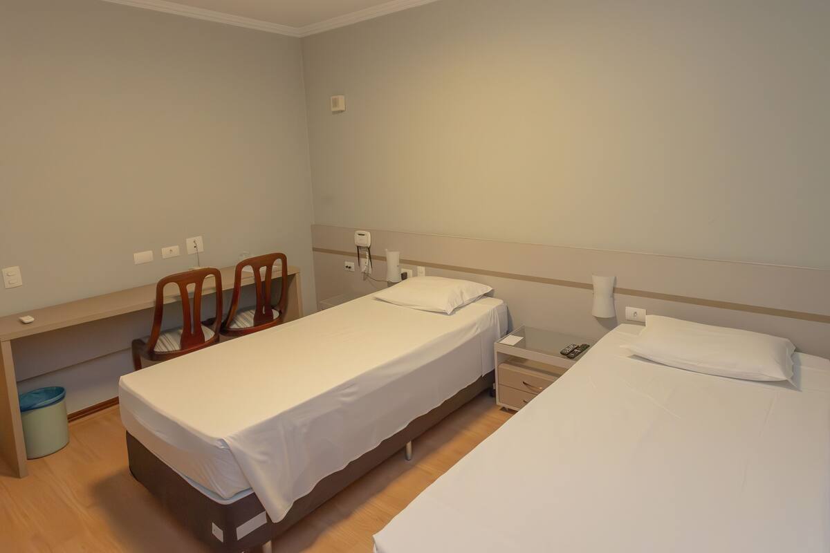 A hotel room with two single beds, a small desk with a chair, a trash bin, and a bedside table with a lamp.