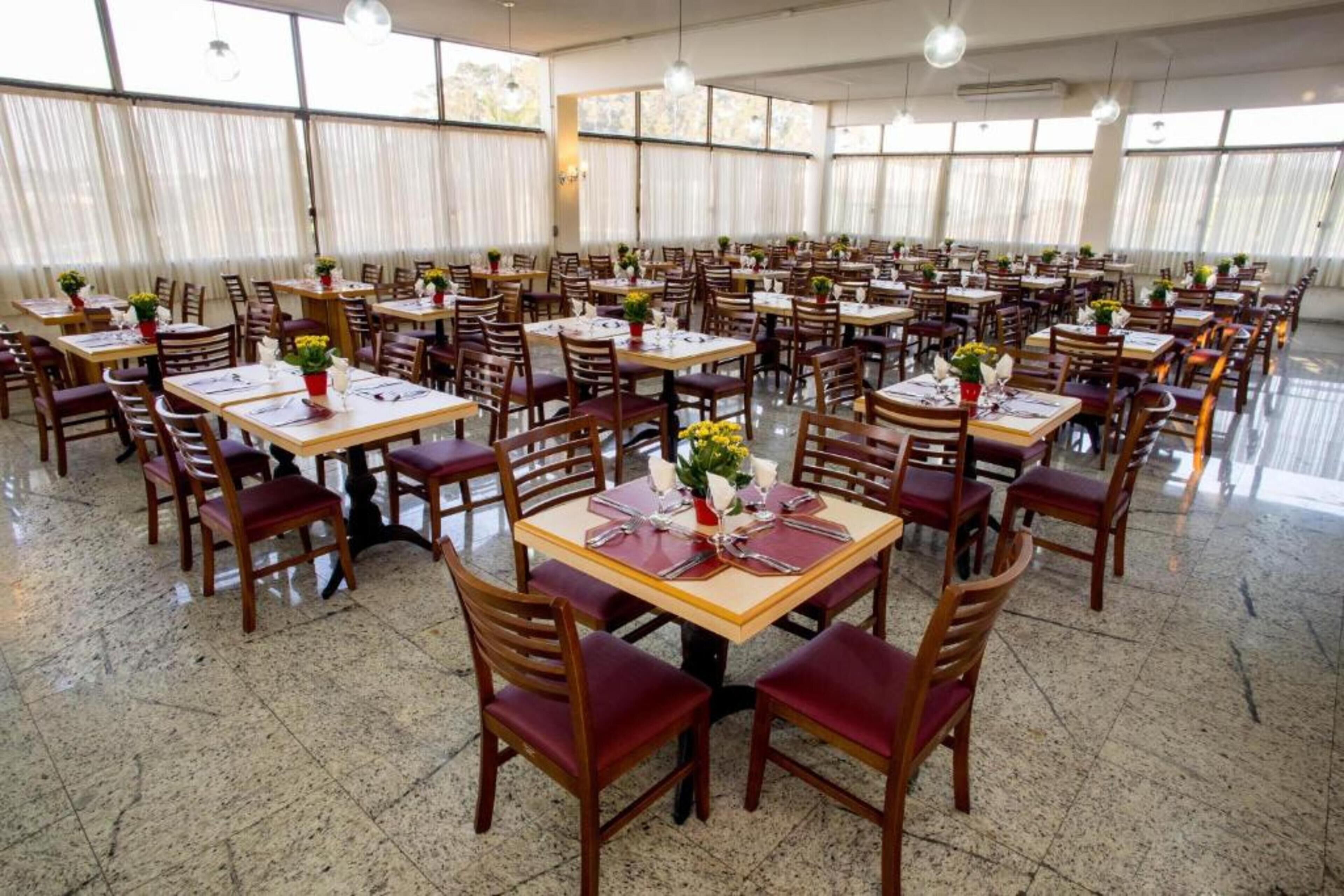 Uma sala de jantar ampla com várias mesas e cadeiras dispostas em fileiras, com grandes janelas que permitem a entrada de luz natural.