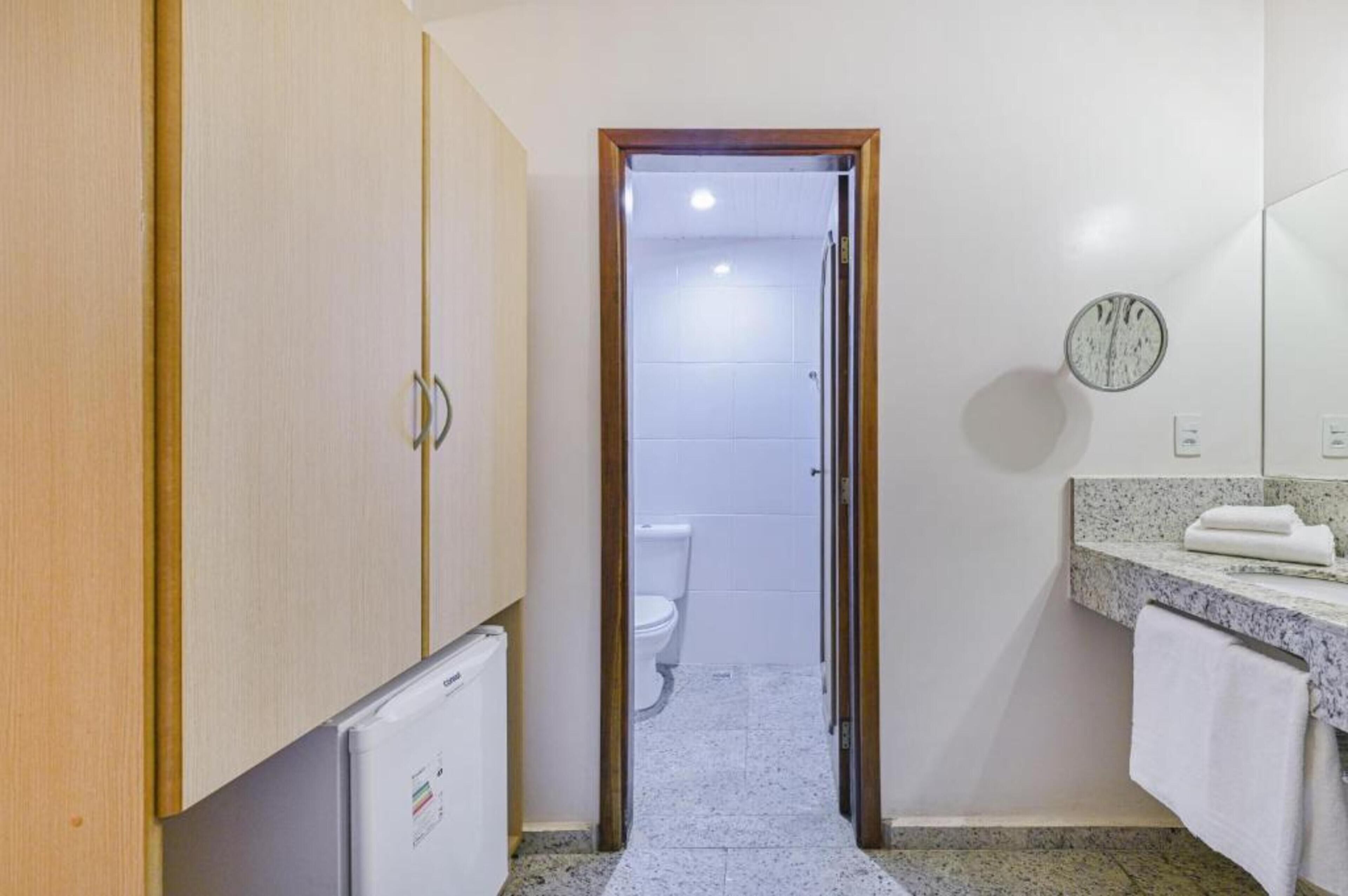 Corredor estreito que dá para um banheiro com vaso sanitário, pia com toalha e relógio na parede.