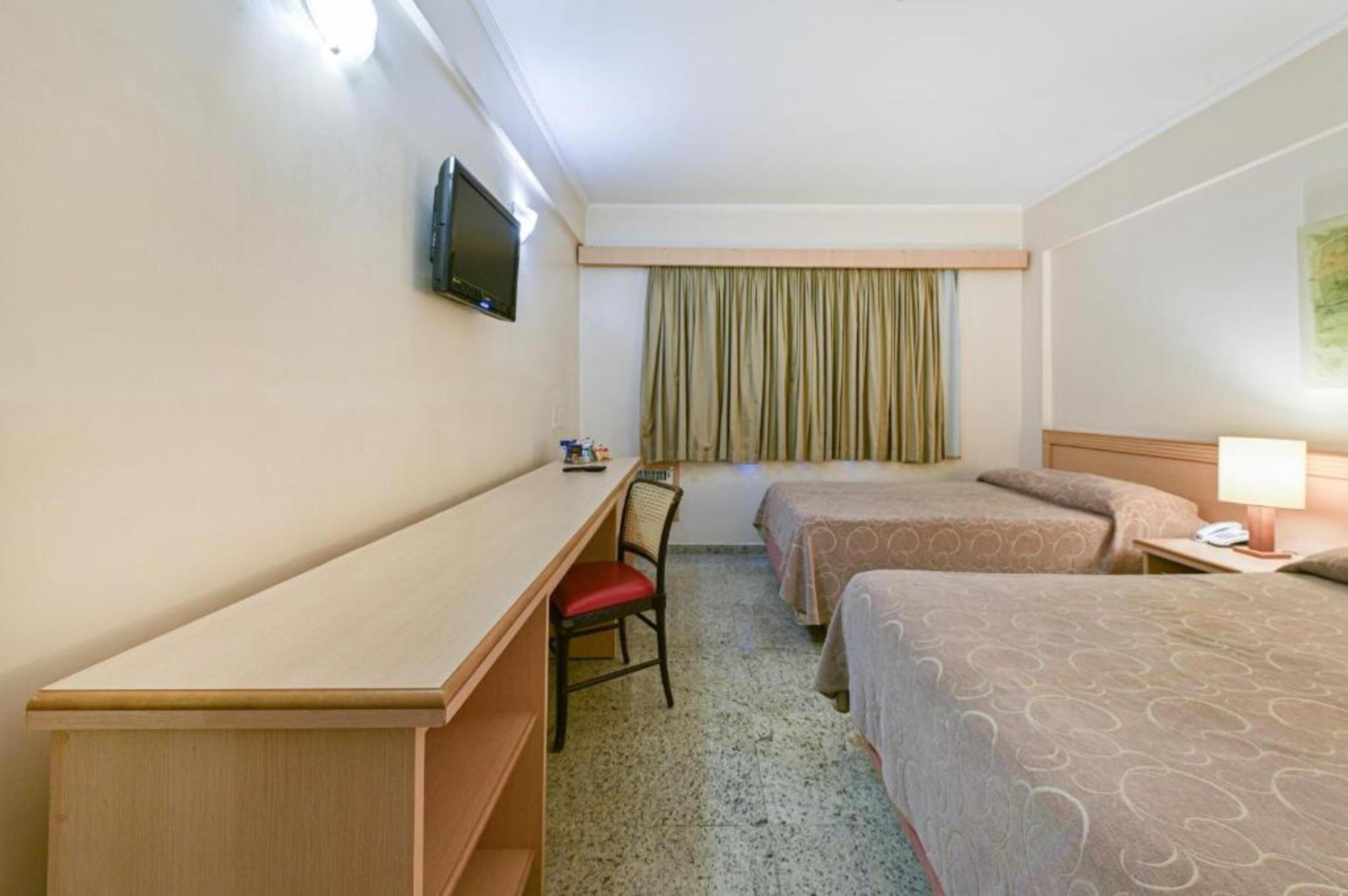 A hotel room with a desk, two beds, a TV, a chair, and a window with curtains.