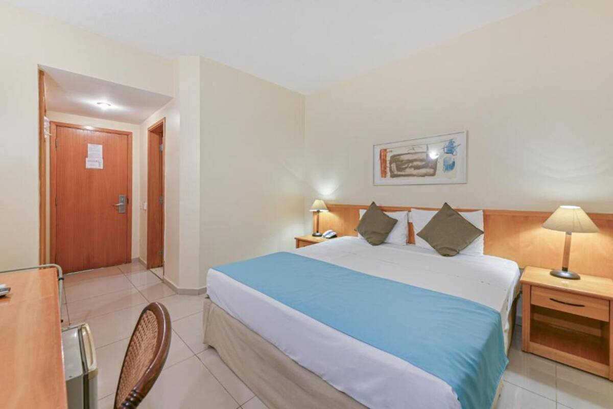 Una habitación de hotel con una cama, mesitas de noche, una silla, un escritorio y una puerta.