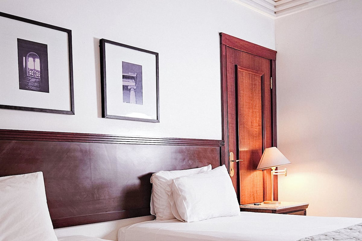 A hotel room with two beds, a wooden headboard, a nightstand with a lamp, and two framed pictures on the wall.