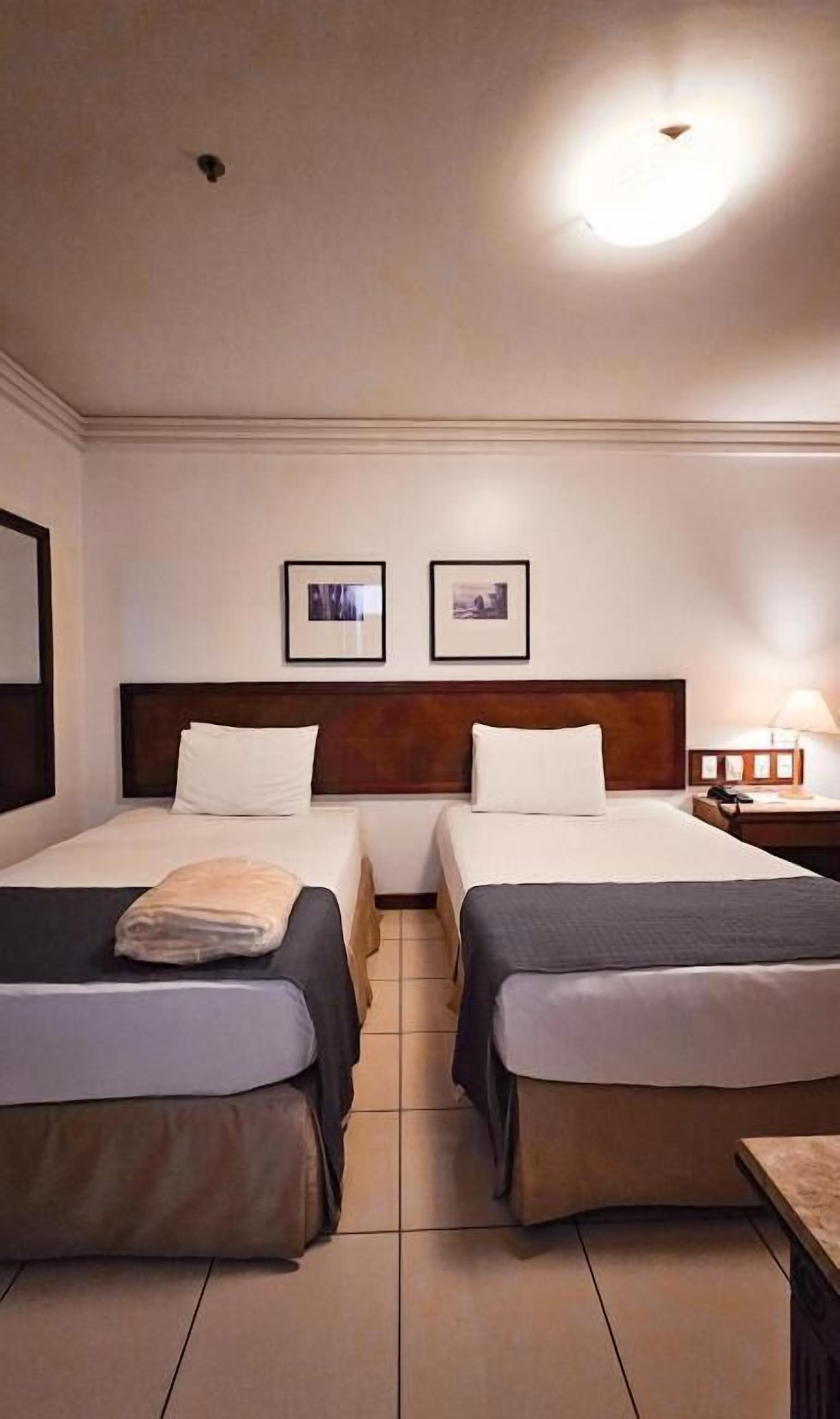 Habitación de hotel con dos camas, cabecera de madera, mesita de noche con lámpara y dos cuadros en la pared.