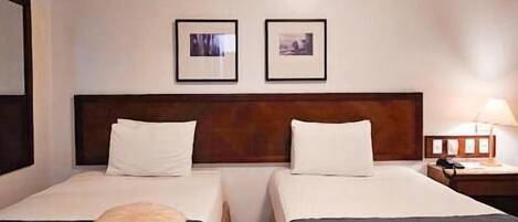 Habitación de hotel con dos camas, cabecera de madera, mesita de noche con lámpara y dos cuadros en la pared.