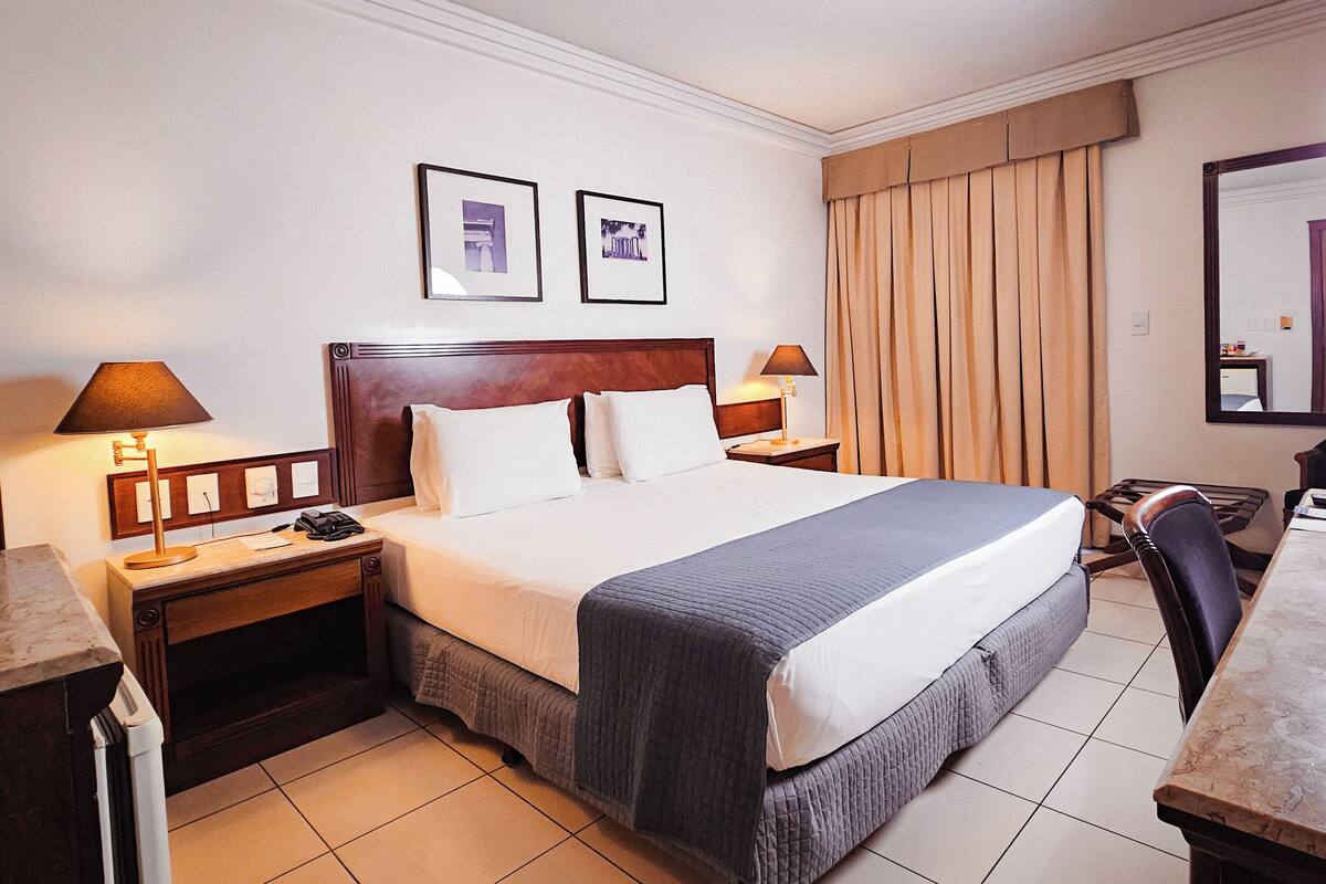 A hotel room with a large bed, two bedside tables, a desk, and a chair. There are two framed pictures on the wall.