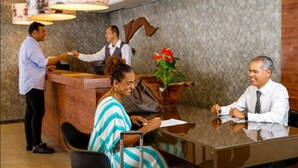 Zona de recepción de un hotel con un recepcionista, un huésped sentado en una mesa y otra persona de pie junto al mostrador.
