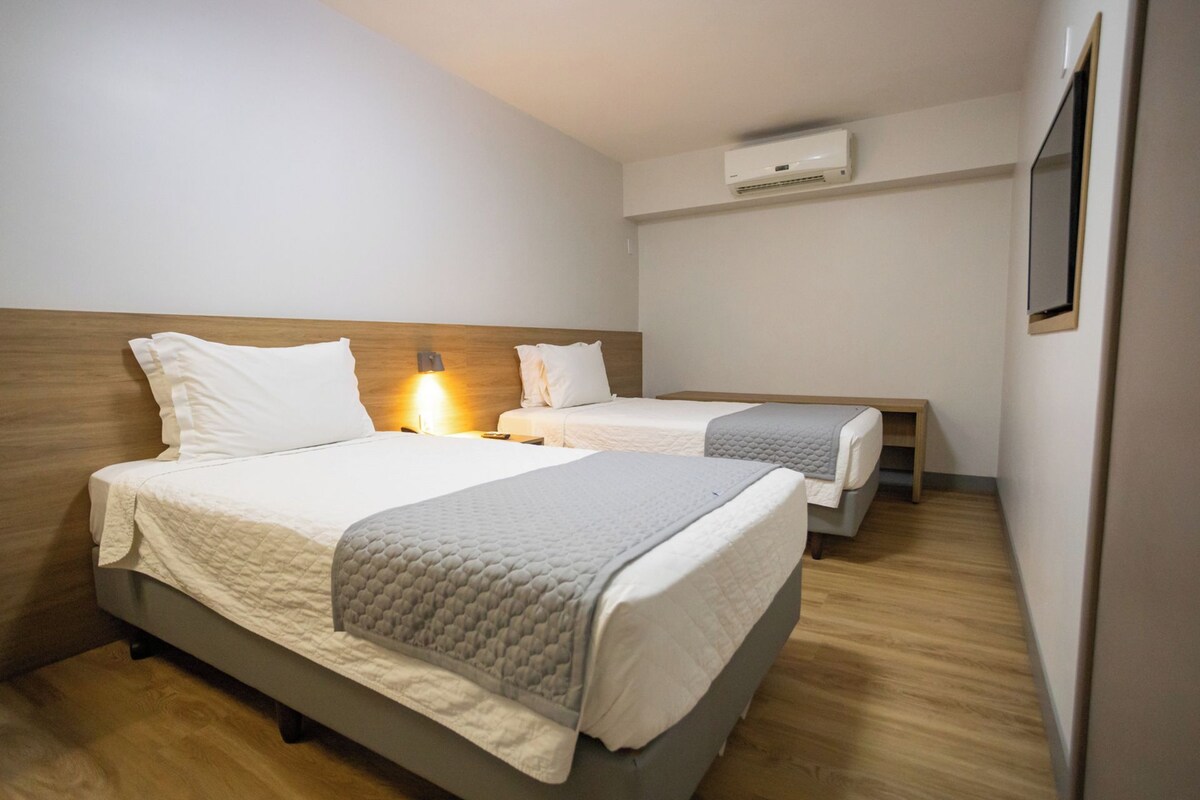 A hotel room with two beds, a bedside lamp, an air conditioning unit, and a television mounted on the wall.